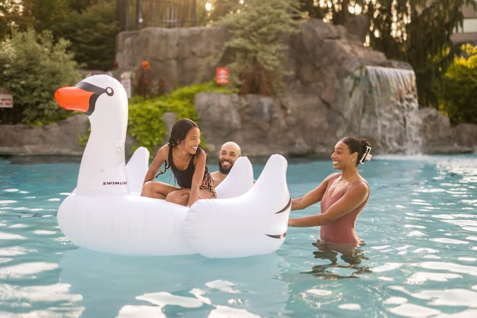 Family enjoying oversized inflatable swan float in resort pool with waterfall feature and stone architecture in background