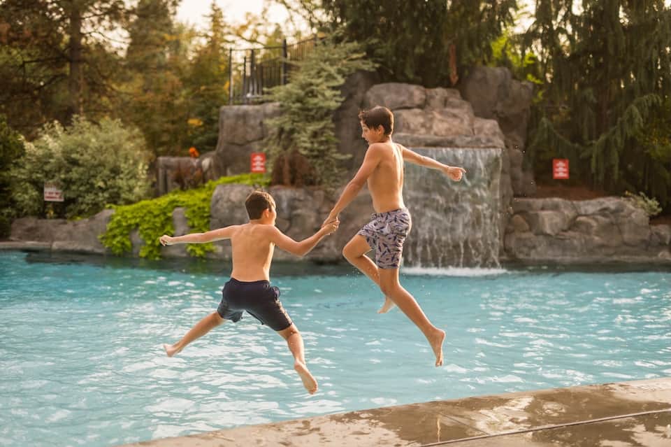Two boys jumping into tropical resort pool with stone waterfall and forest backdrop