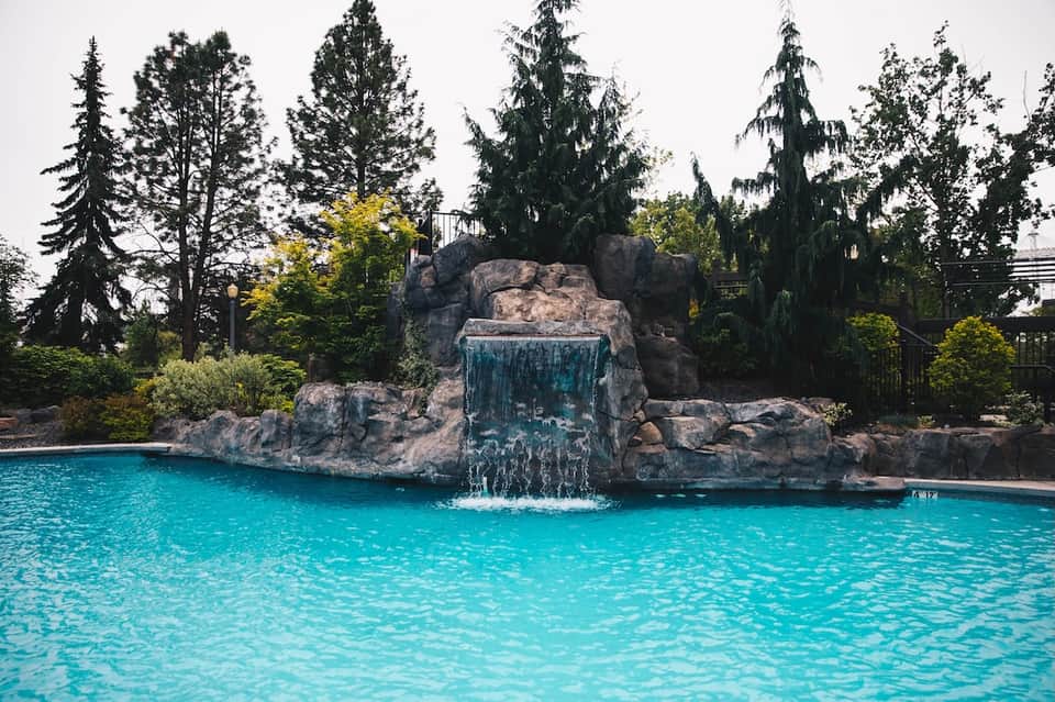 Turquoise pool with waterfall cascading from large rock formation surrounded by tall evergreen trees