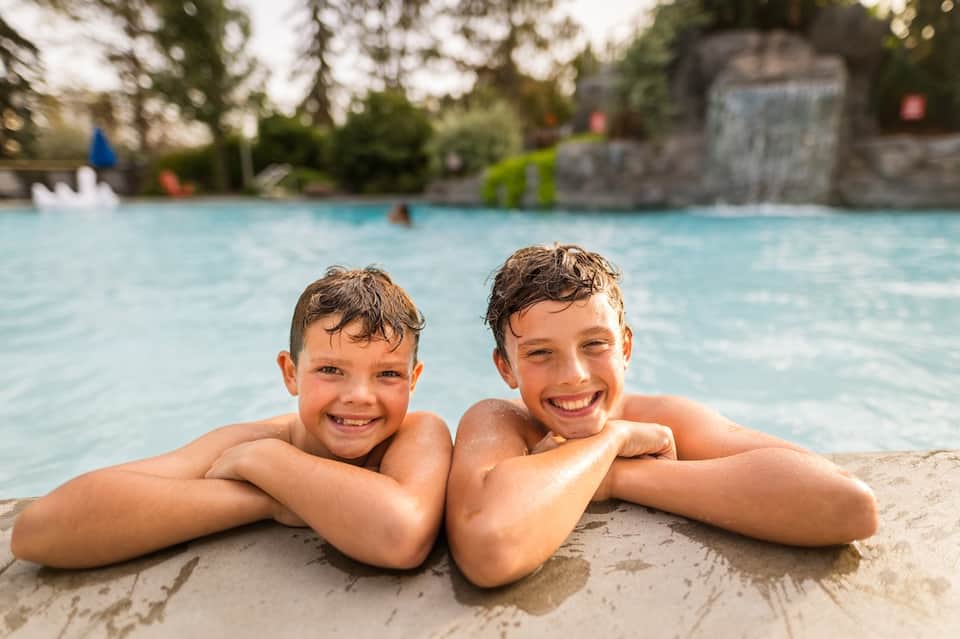 Two children smiling in resort pool with water slide and stone structure in background