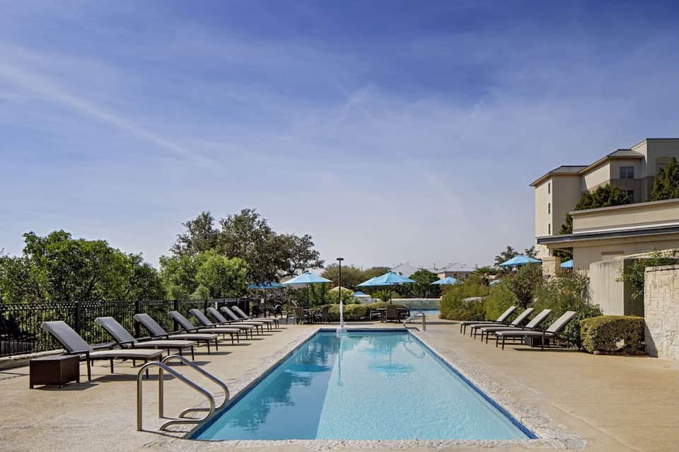 Resort pool with loungers, blue umbrellas, modern building, manicured landscaping, and clear blue sky