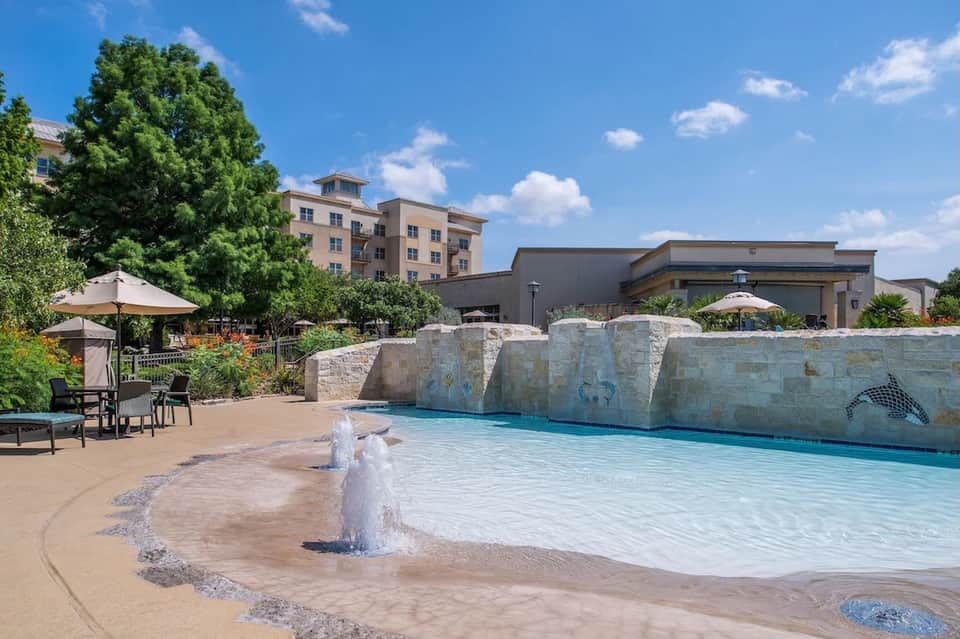 Resort pool with wave features and stone waterfall backdrop, surrounded by umbrellas and hotel buildings