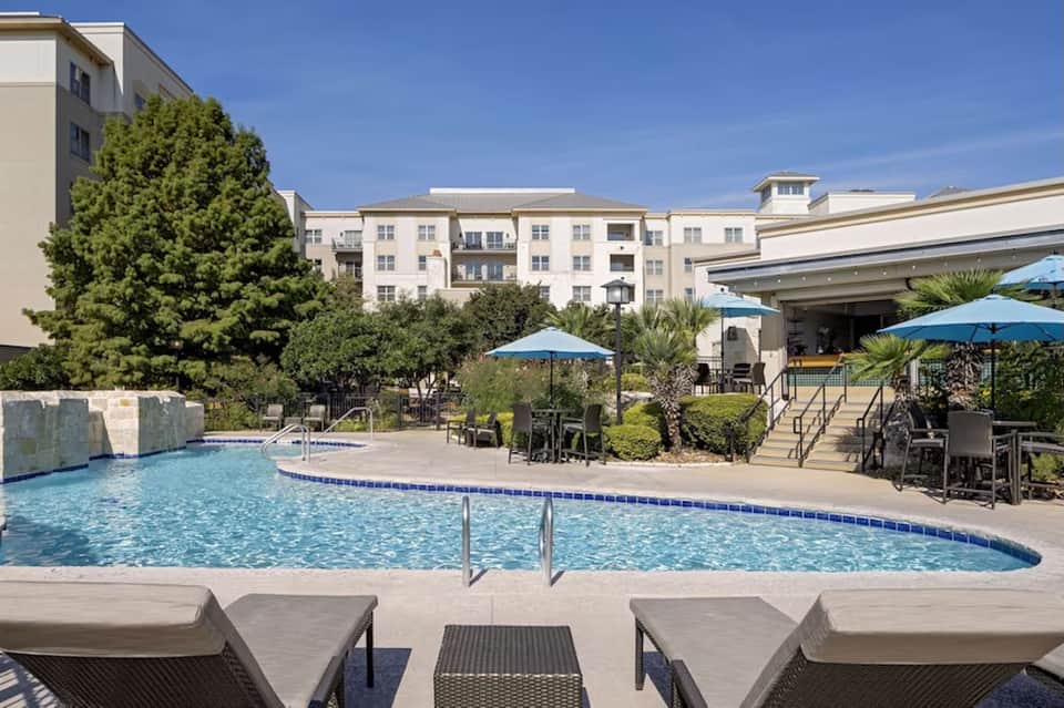 Resort pool area with lounge chairs, blue umbrellas, landscaping, and multi-story buildings