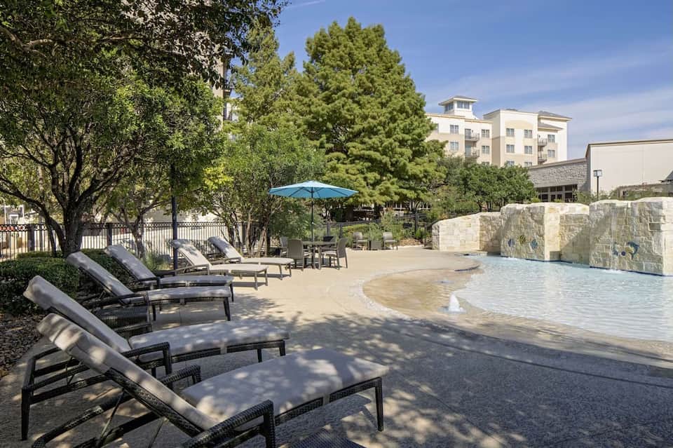 Lazy river resort area with lounge chairs, blue umbrella, natural landscaping, and hotel building backdrop