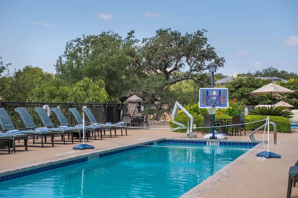 Resort pool with basketball hoop, loungers under blue umbrellas, and mature oak trees in background