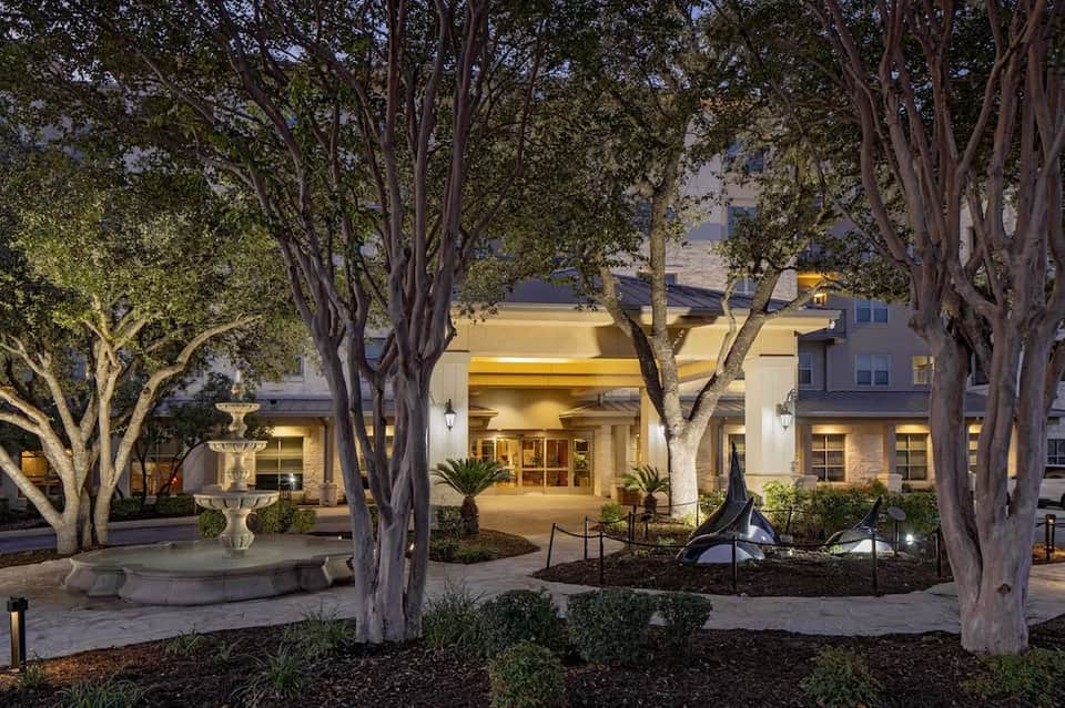 Hotel entrance at dusk with tall trees, fountain, and illuminated glass doors