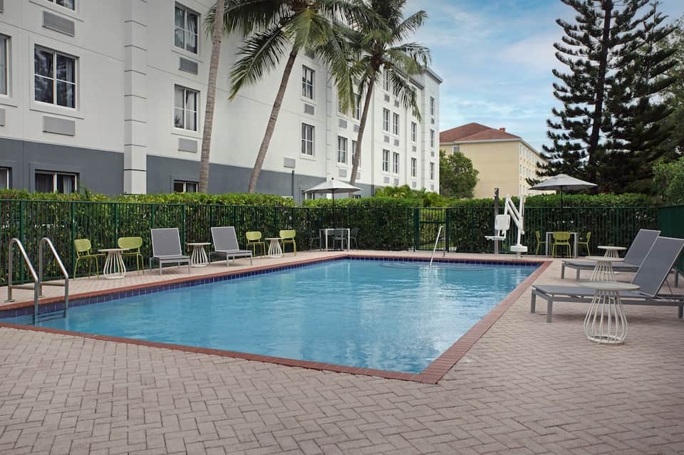 Resort rectangular pool with brick surround, loungers, high-rise hotel building backdrop, and palm trees lining fence