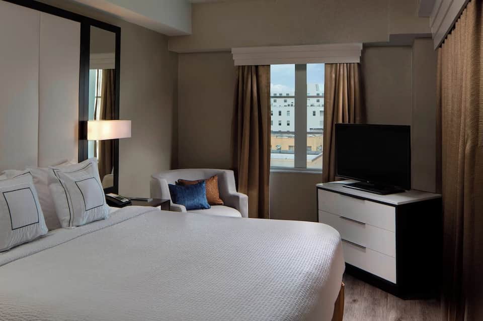 Modern hotel bedroom with king bed, lounge chair, flat-screen TV, and city skyline view