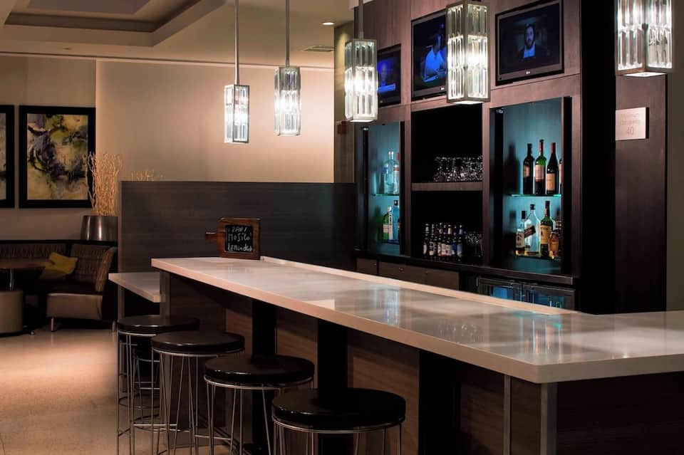 Upscale hotel bar with dark wood cabinetry, illuminated shelving displaying spirits, and modern pendant lighting