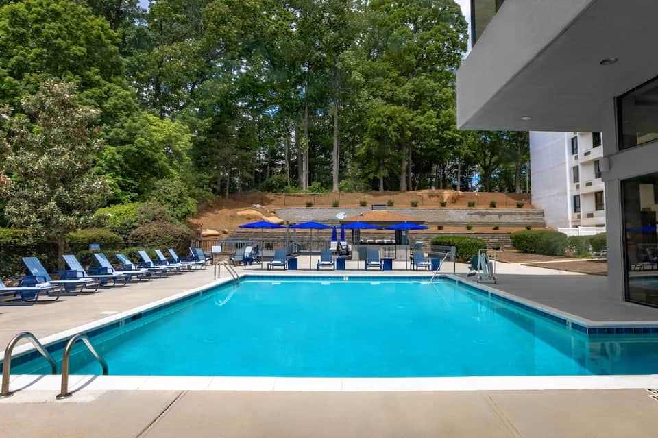 Blue pool with blue lounge chairs and umbrellas backed by modern building and trees