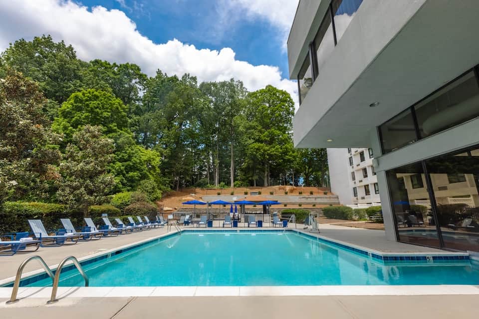 Resort pool surrounded by blue loungers, trees, and modern white building with sky