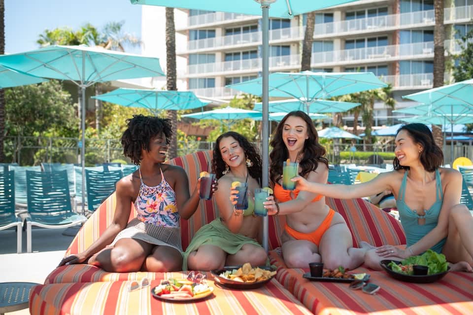 Resort pool deck with turquoise umbrellas, striped loungers, diverse group of guests with drinks, and building backdrop