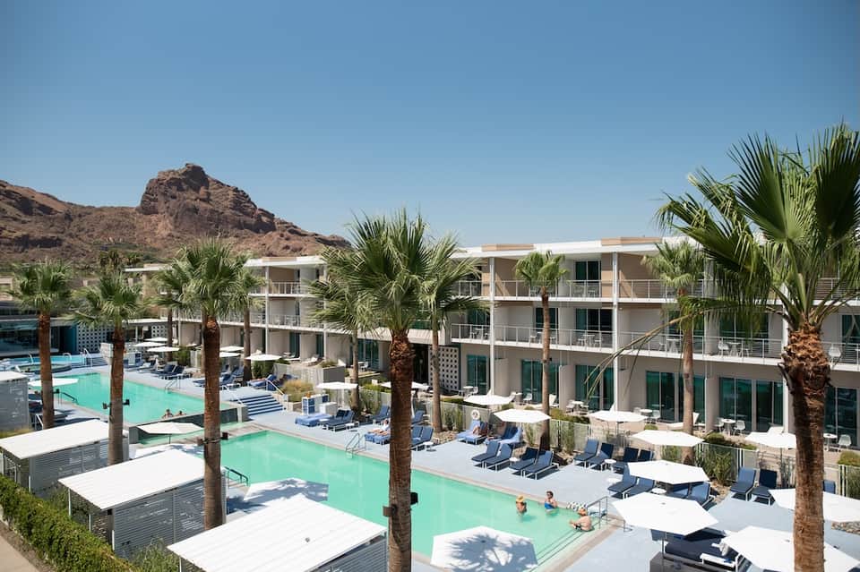 Resort aerial view with turquoise pools, white loungers, palm trees, and desert mountain backdrop