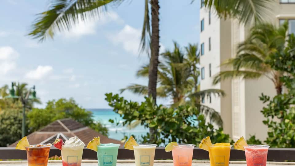 Array of colorful tropical cocktails with citrus garnishes displayed against ocean and palm tree backdrop