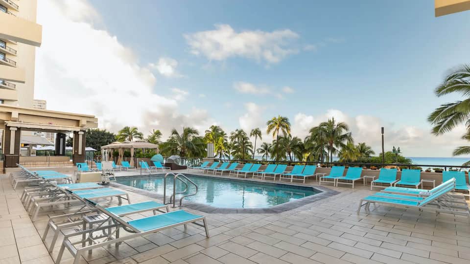 Resort pool deck with turquoise lounge chairs, umbrellas, palm trees, and ocean view
