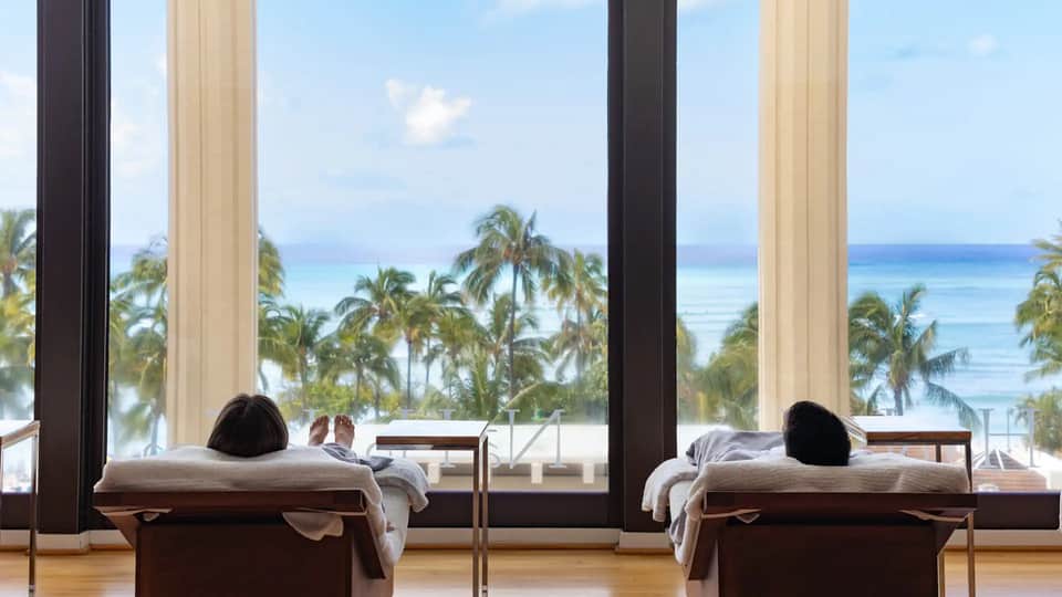 Spa treatment room with massage beds overlooking ocean and palm trees through large windows