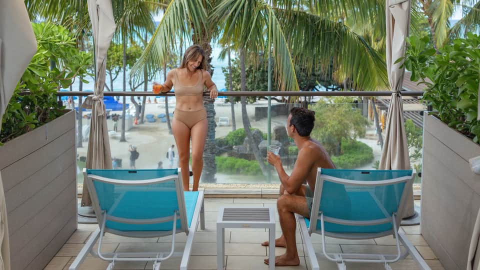 Private balcony with couple in swimwear, turquoise lounge chairs, palm trees, and ocean view