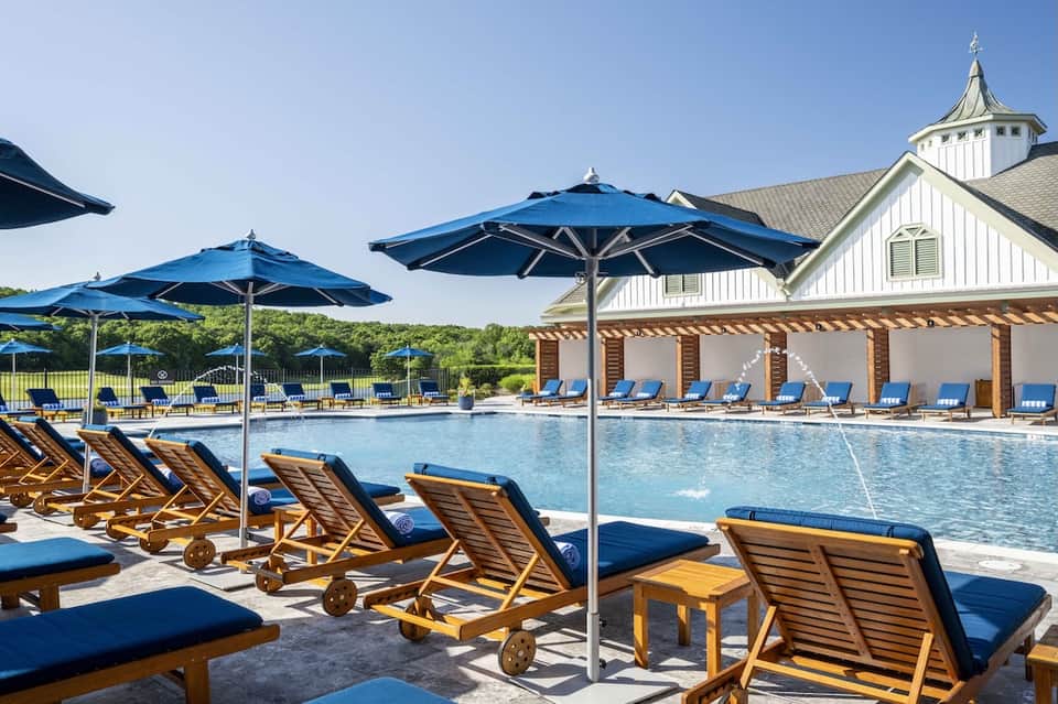 Resort pool with blue lounge chairs, umbrellas, white clubhouse, and manicured grounds