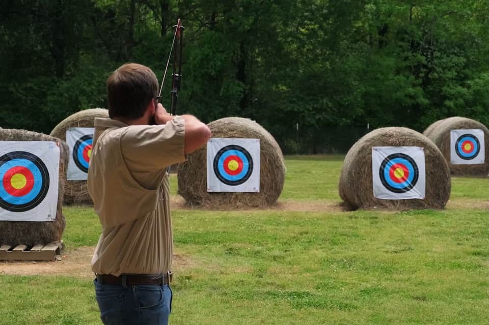 Archer drawing compound bow at outdoor range with hay bale targets in forest setting