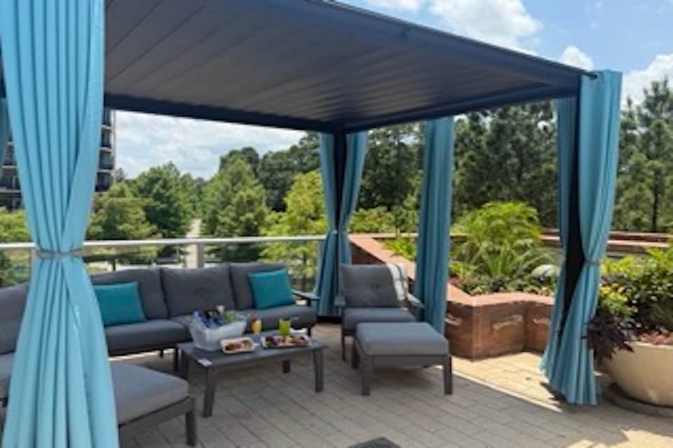 Covered rooftop lounge with pergola, gray outdoor seating, teal cushions, and green forest views