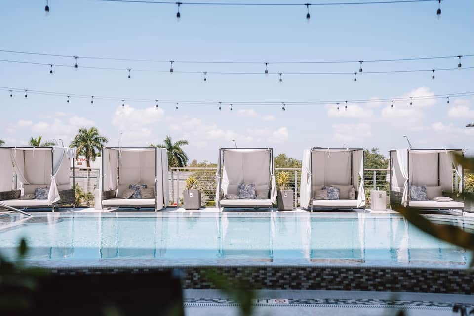 Luxury resort infinity pool with white cabanas and string lights under blue sky with palm trees.
