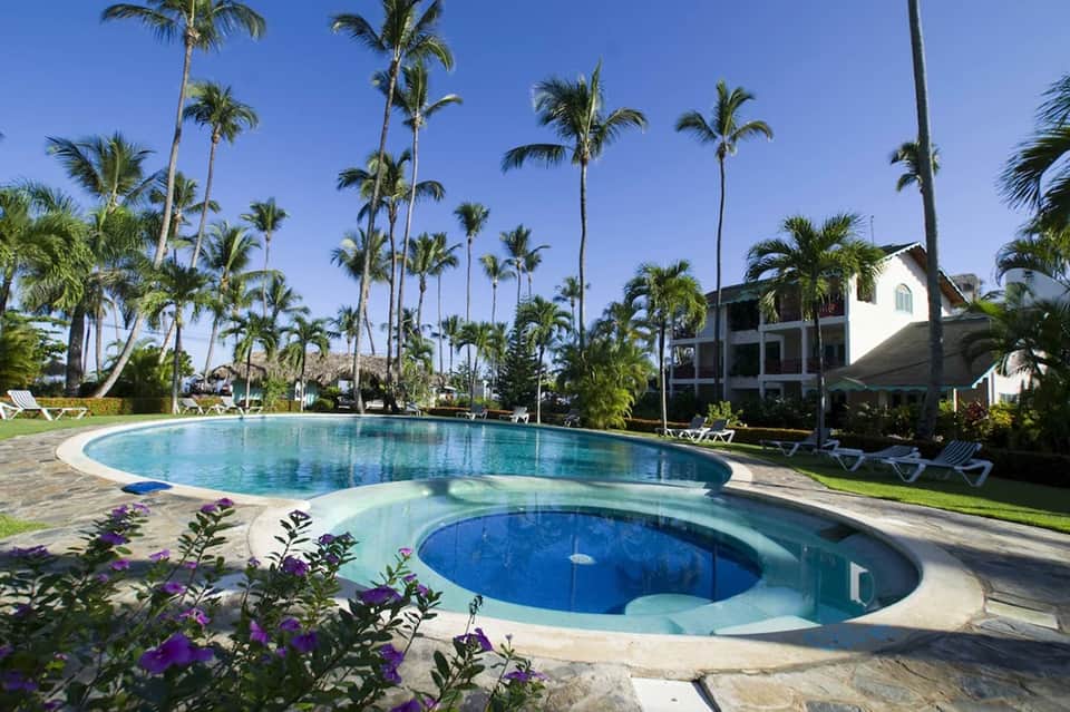 Resort pool complex with palm trees, lounge chairs, and white villa-style buildings