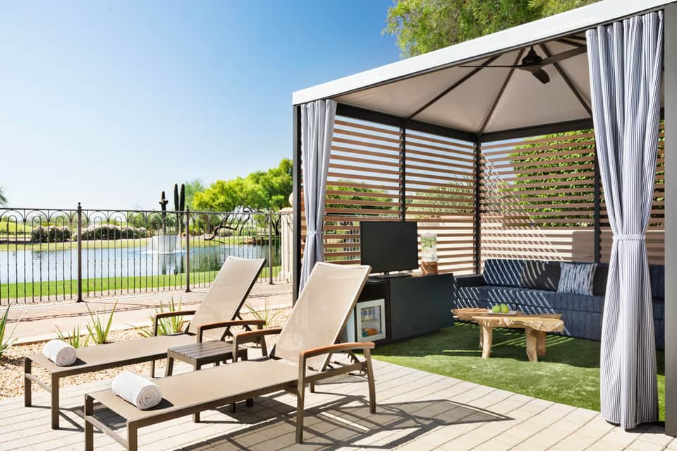 Modern poolside gazebo with striped lounge chairs, fire pit, and water views behind metal fence