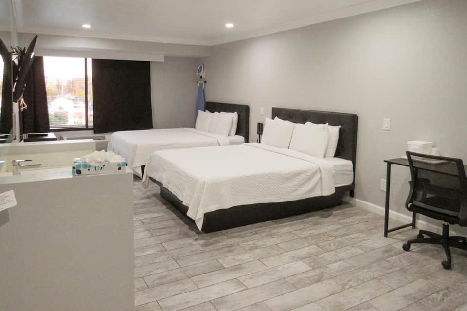 Modern hotel room with two queen beds, black headboards, white bedding, desk area, and bathroom