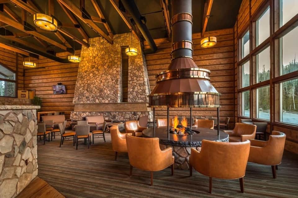 Rustic lodge interior with wooden beams, stone fireplace, central fire pit, and leather armchairs