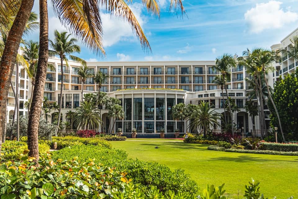 Palm tree-lined courtyard of white hotel building with manicured lawn and clear blue sky