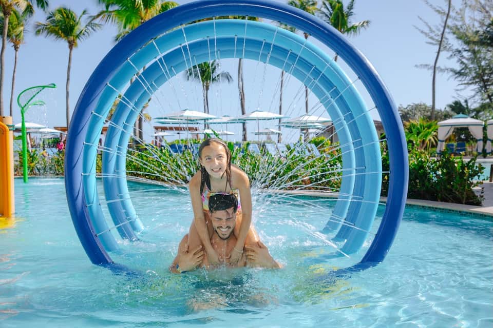 Father and daughter playing in pool through large blue tunnel toy with palm trees and umbrellas