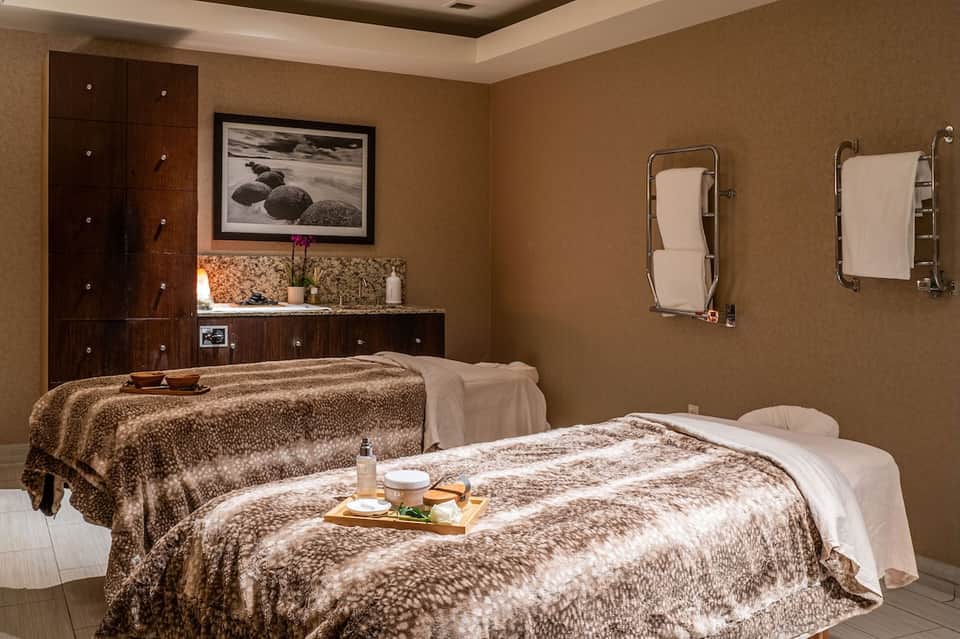 Spa treatment room with two massage tables, warm earth tones, and dark wood cabinetry