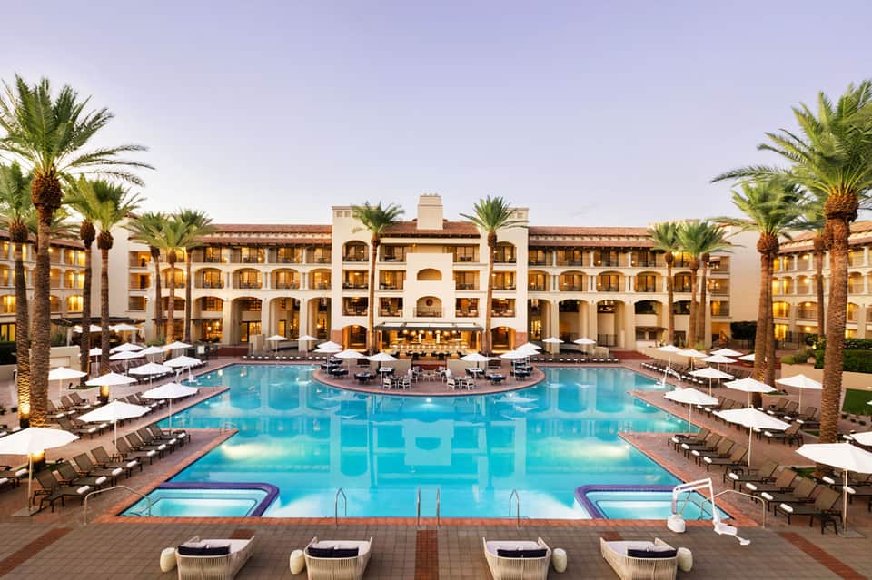 Large resort pool surrounded by Mediterranean-style buildings with palm trees and white loungers