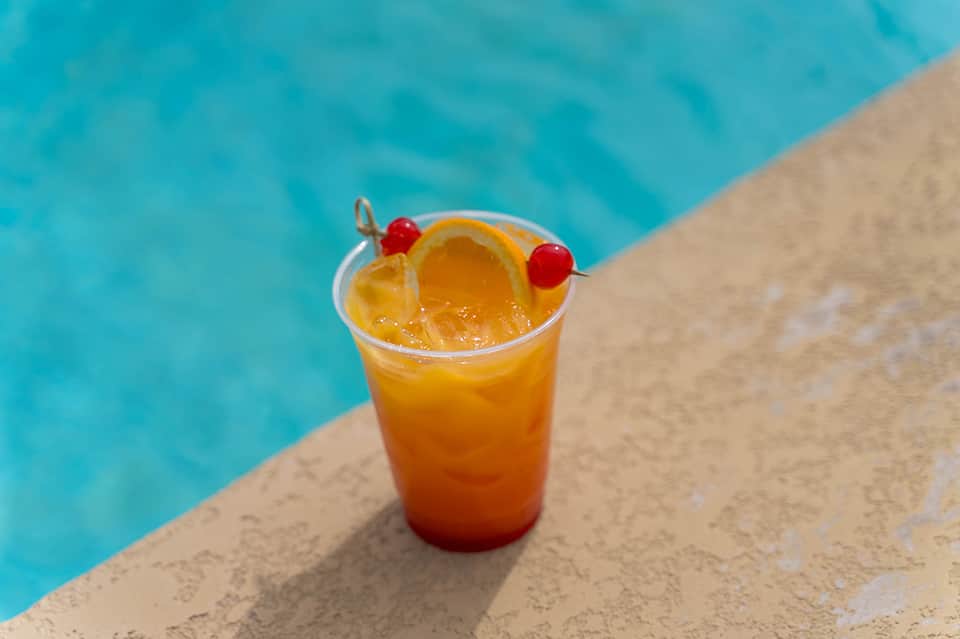 Orange tropical cocktail with orange slice and cherries on pool deck beside turquoise water