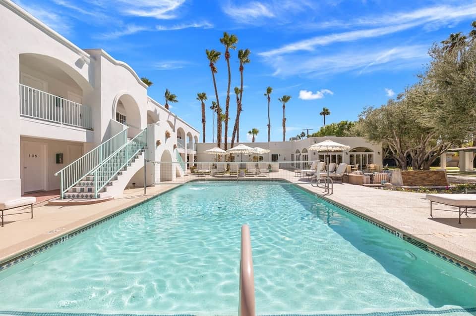 Rectangular pool with white umbrellas, palm trees, modern white building, and desert landscape