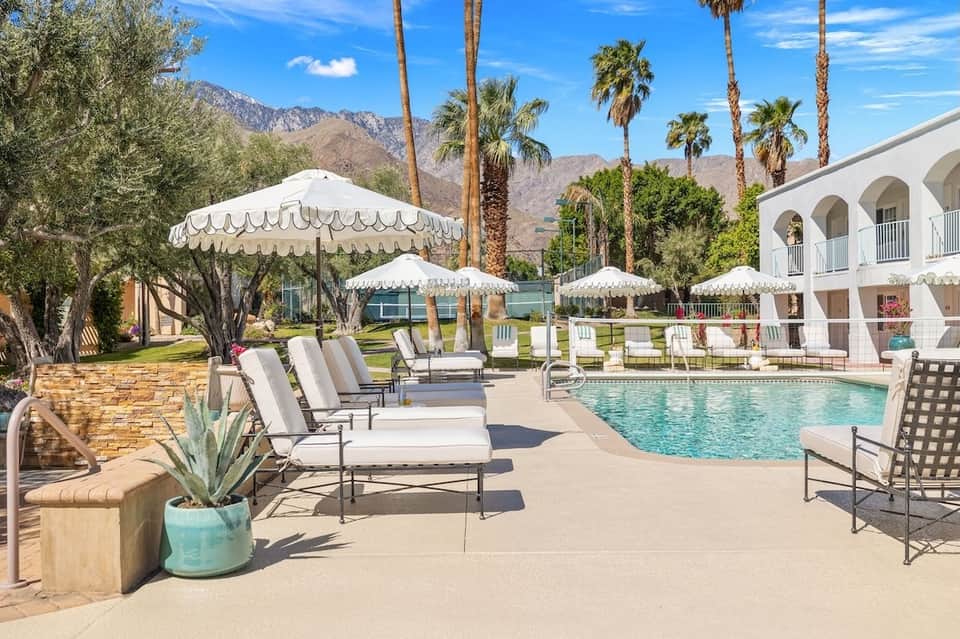 Resort pool area with white umbrellas, lounge chairs, palm trees, and mountain backdrop