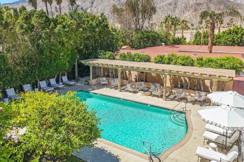 Desert resort pool with wooden pergola shade structure, lounge chairs, mountains in background, and lush landscaping