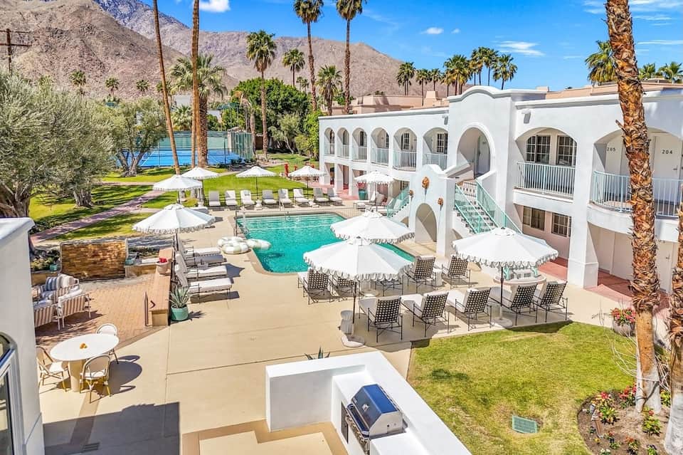 Pink and white Moorish-style resort with turquoise pool, white umbrellas, palm trees, and desert mountains
