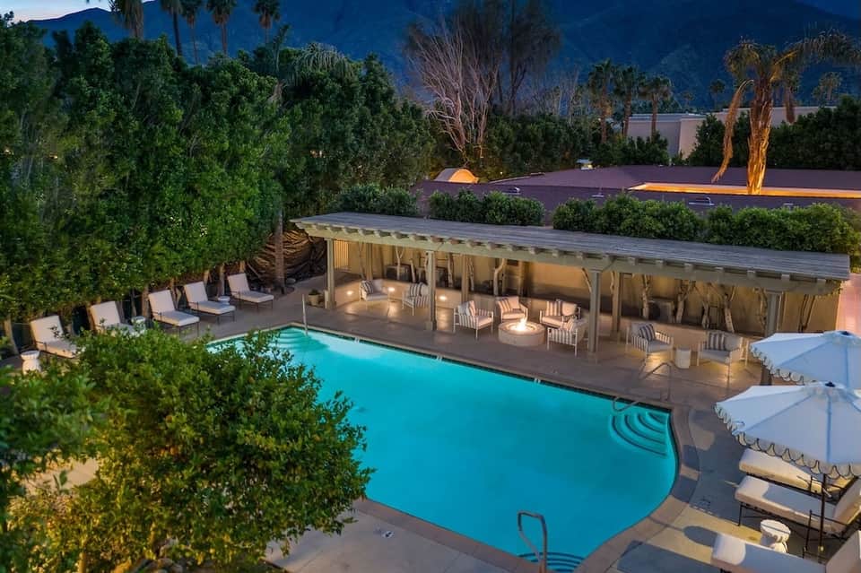 Luxury desert resort pool at night with cabana pavilion, teak lounge chairs, illuminated eucalyptus tree, and mountains.