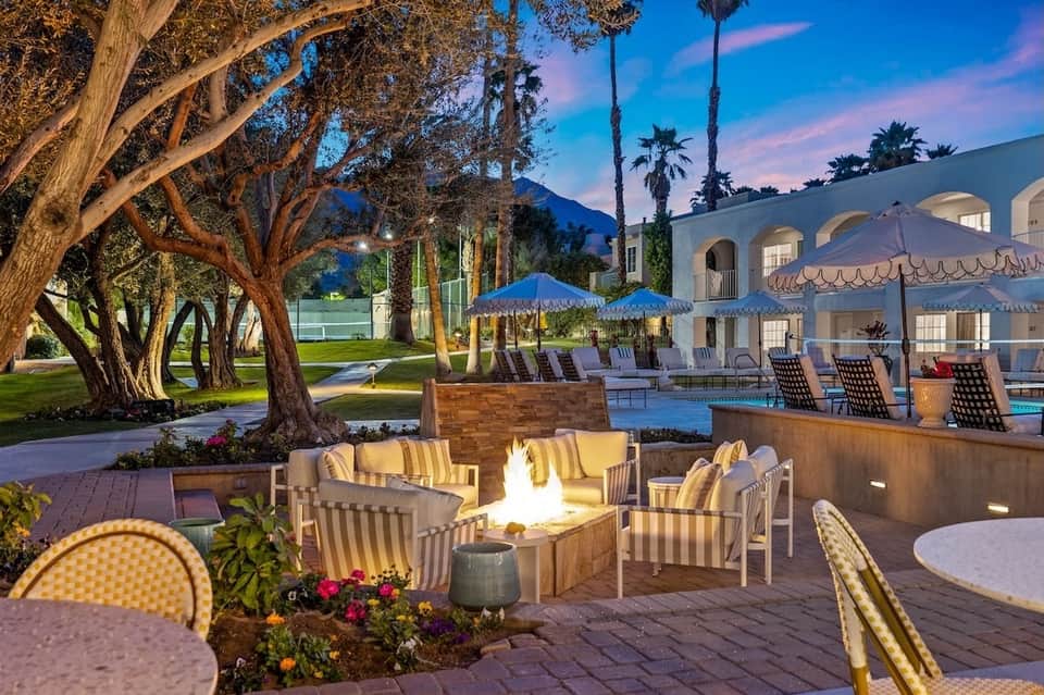 Evening outdoor fireplace seating area with striped chairs, fire pit, palm trees, and lit building in background