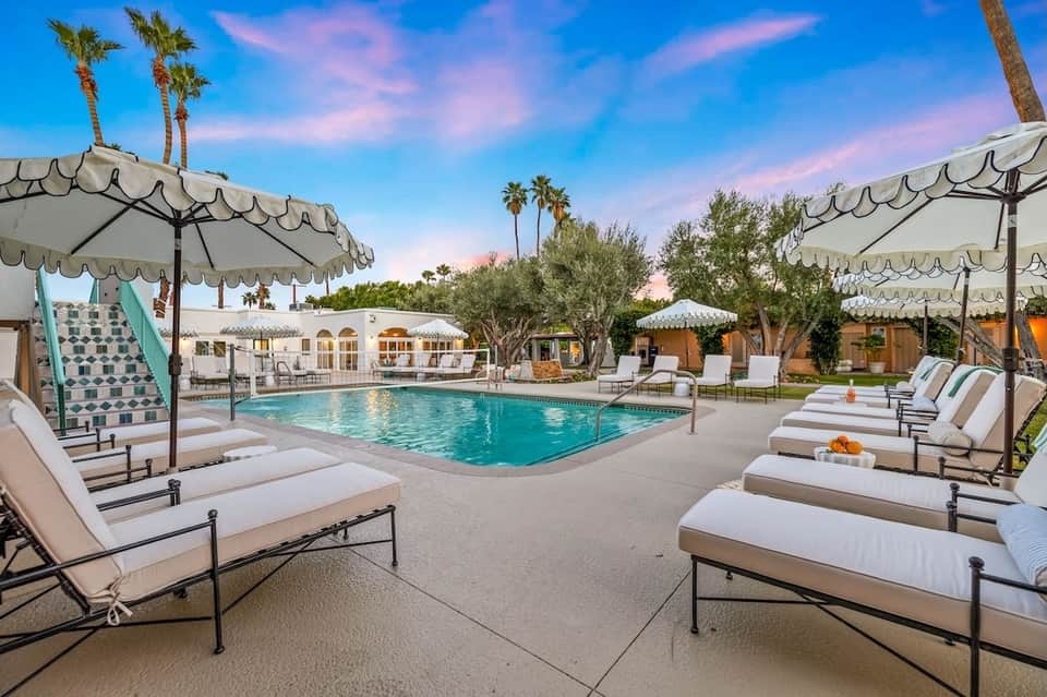 Upscale resort pool at dusk with umbrella-shaded loungers, palm trees, and white buildings in background