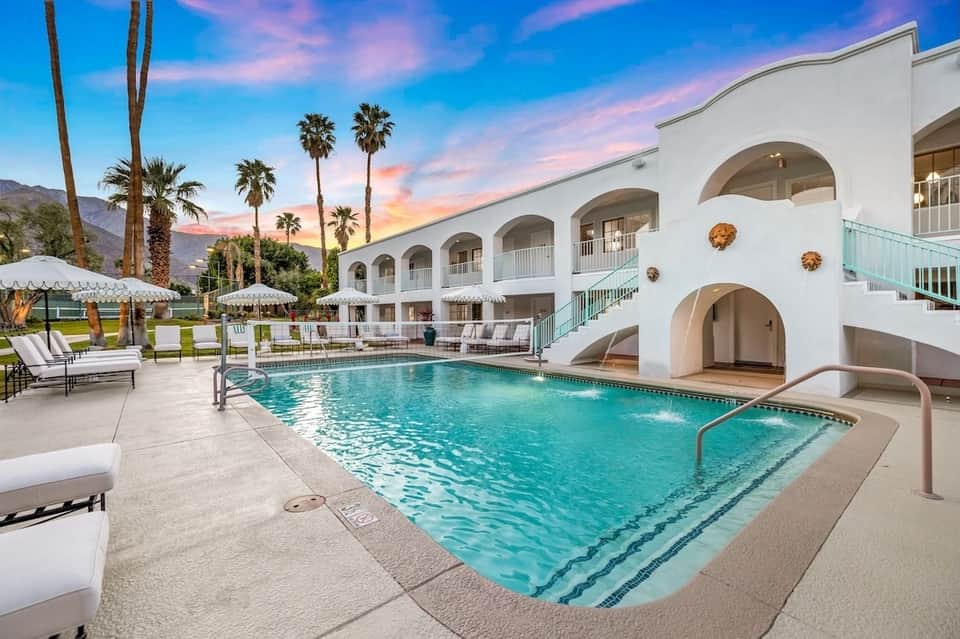 Resort pool with lounge chairs, umbrellas, palm trees, and white arched building at sunset