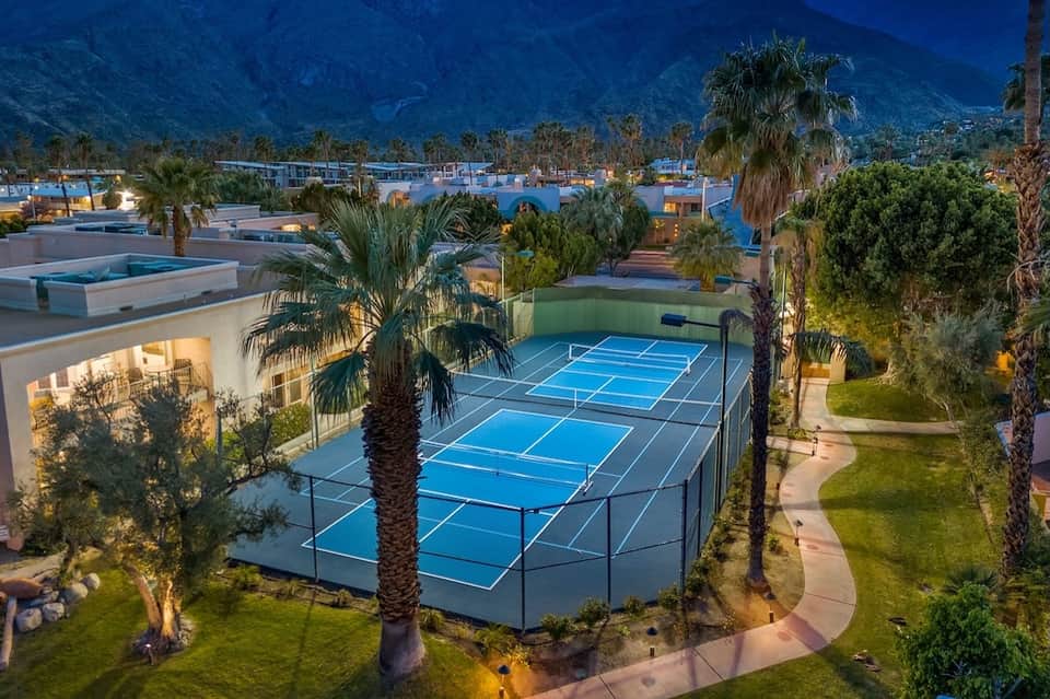 Aerial view of resort with multiple pools, tennis courts, palm trees, and evening lighting