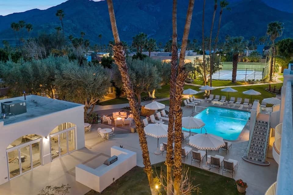 Luxury resort grounds at dusk with illuminated pools, palm trees, mountain backdrop, and loungers