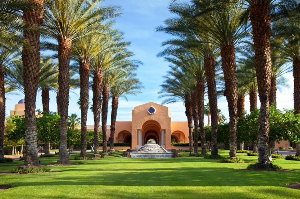 Luxury resort courtyard with palm-lined pathway leading to terracotta building with arched entrance and manicured lawn