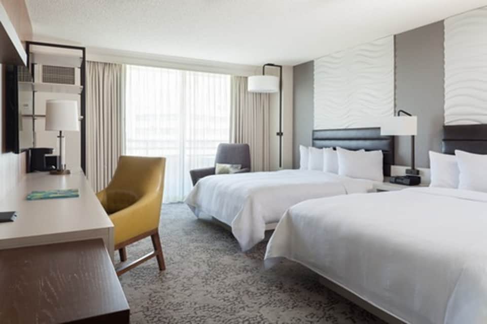 Modern hotel room with two beds, seating area, large windows, and neutral tones