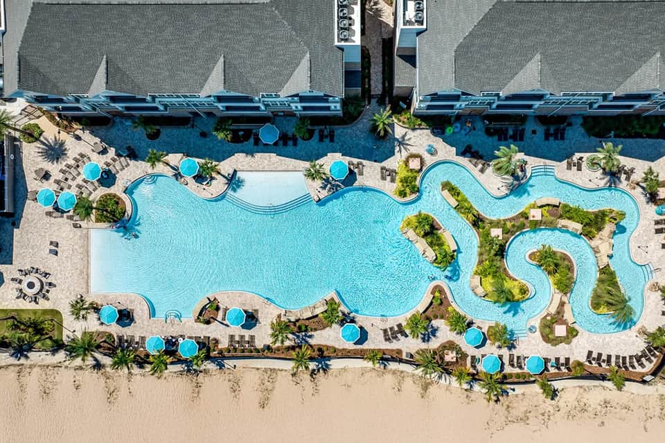 Aerial view of curved resort pool with blue umbrellas and lounge chairs along the beach