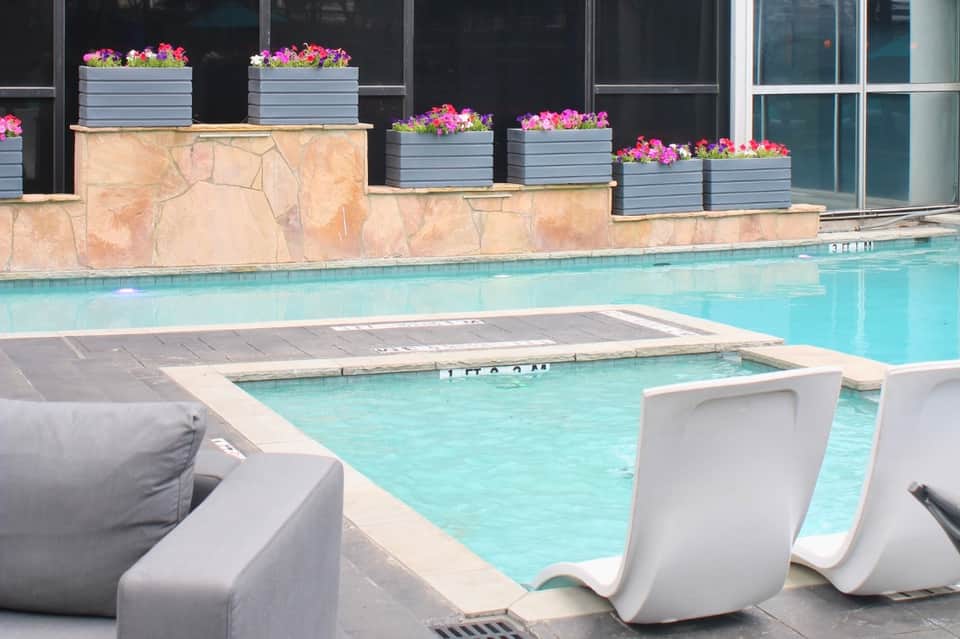 Resort pool with heated spa, pink flowers in planters, and lounge chairs against glass building