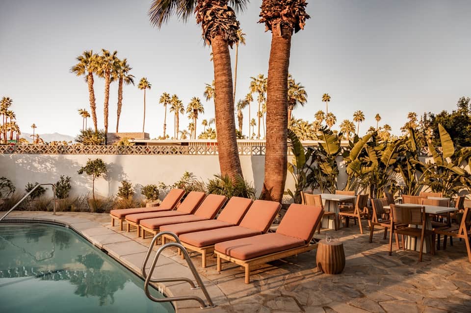 Resort poolside lounge area with orange cushioned chairs, tall palm trees, and white walls