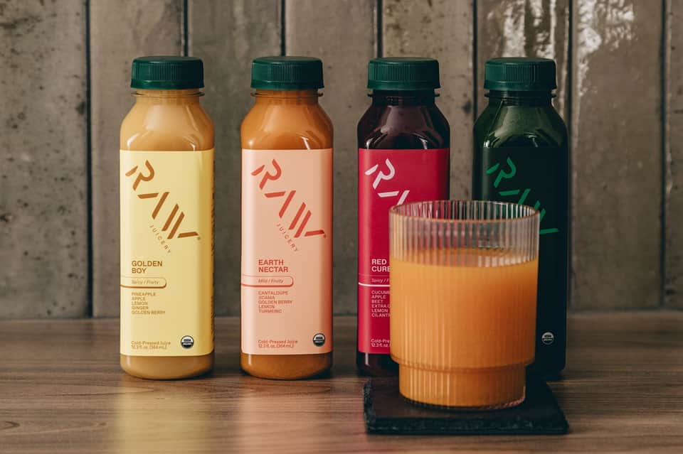 Four colorful cold-pressed juice bottles with green caps displayed with a full glass